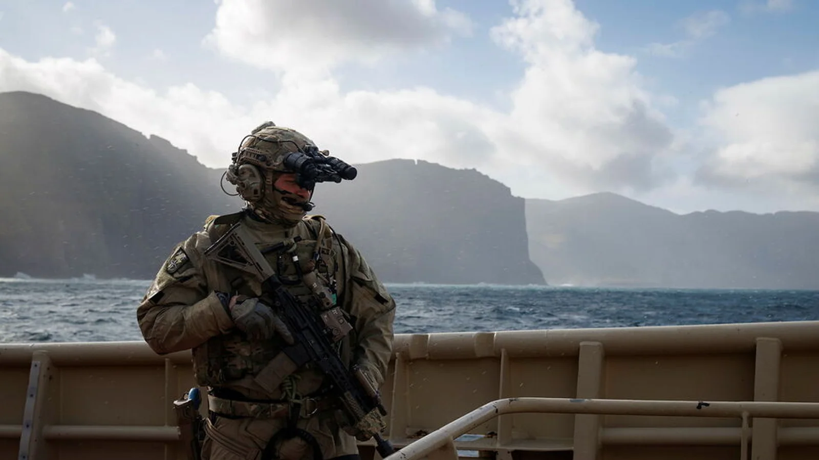 Royal Marine aboard a vessel with steep coastal cliffs behind him during Exercise Lion Protector 26