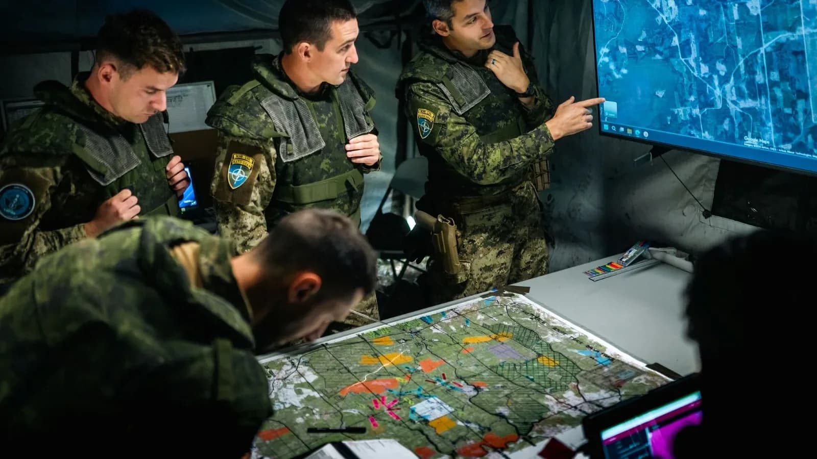 Military personnel discuss maps and a digital display in a command setting
